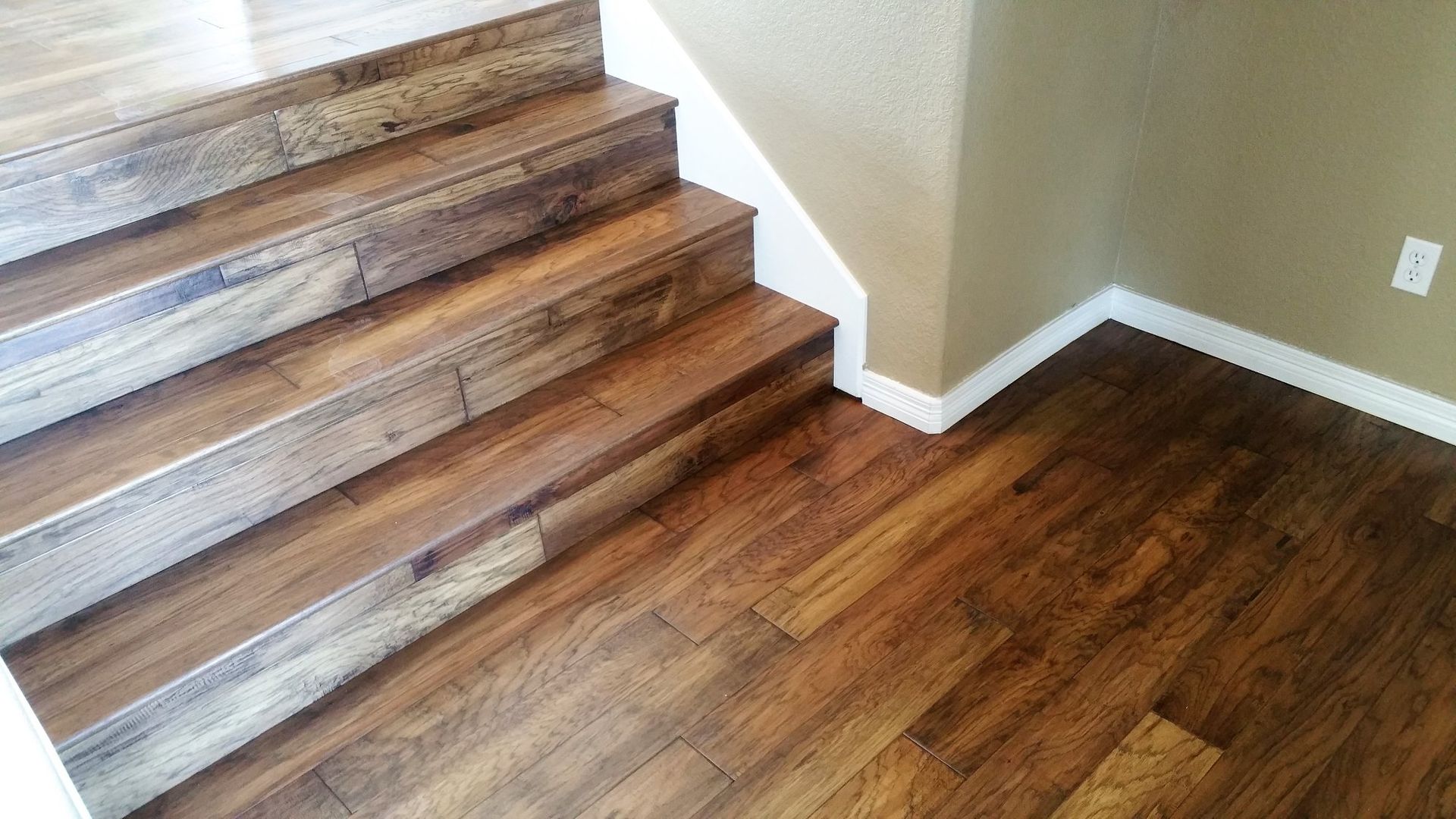 Wooden stairs and flooring with light and dark brown tones, next to a tan wall and white trim.