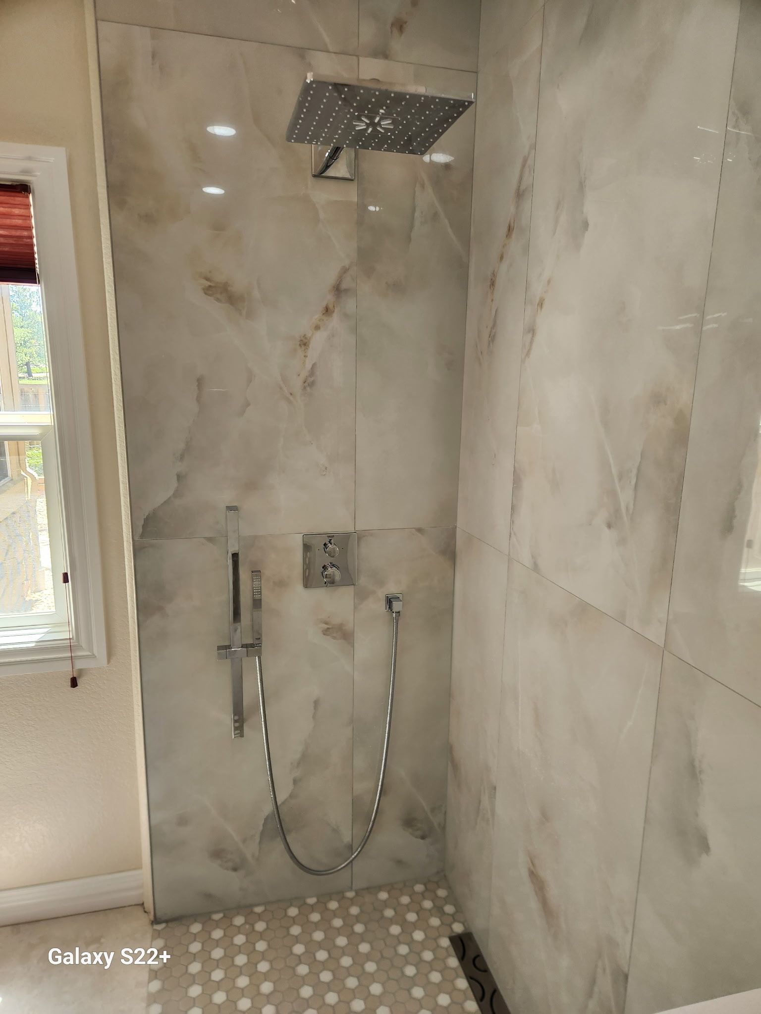 Modern tiled shower with rainfall head, handheld sprayer, and mosaic floor next to a window.