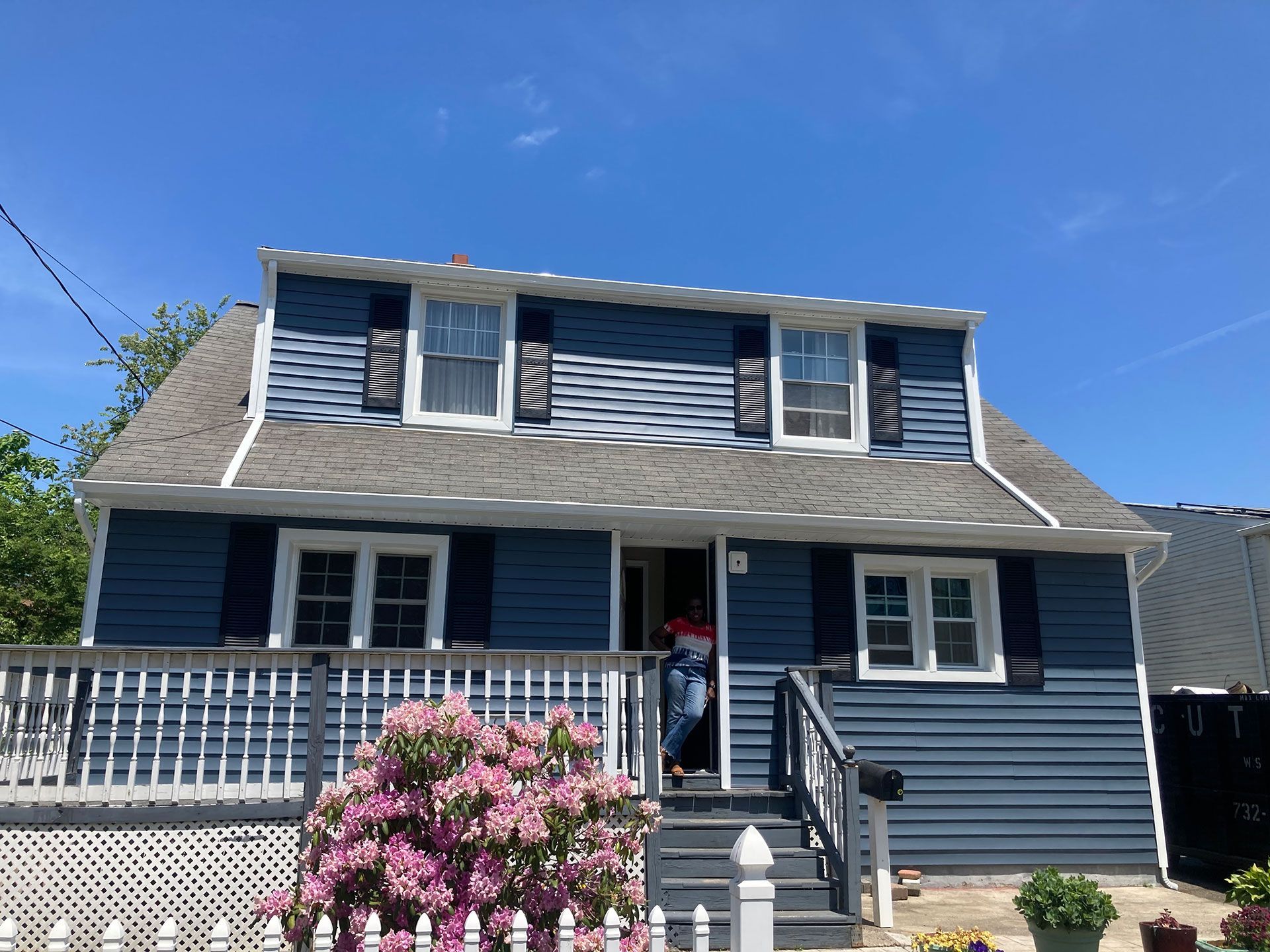 A blue house with white shutters and a pink bush in front of it.