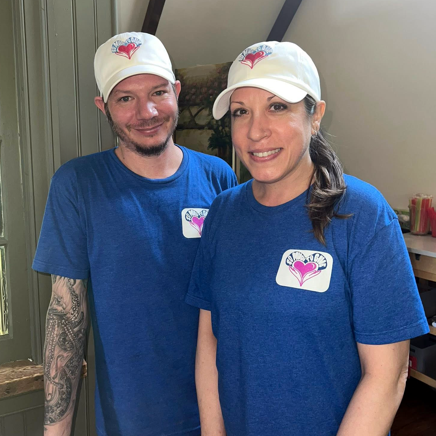 Two people wearing matching blue shirts and white hats with a logo, smiling indoors.