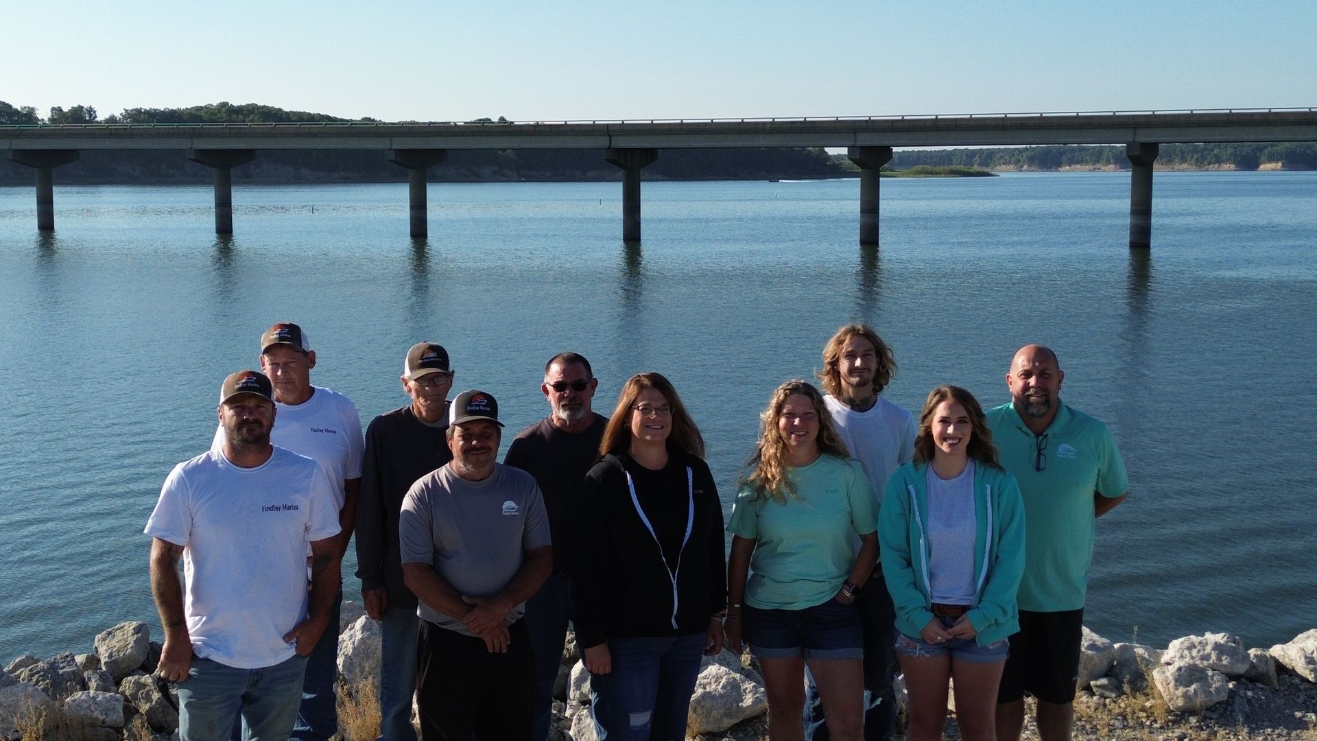 Group of people standing by a lake in front of a bridge.