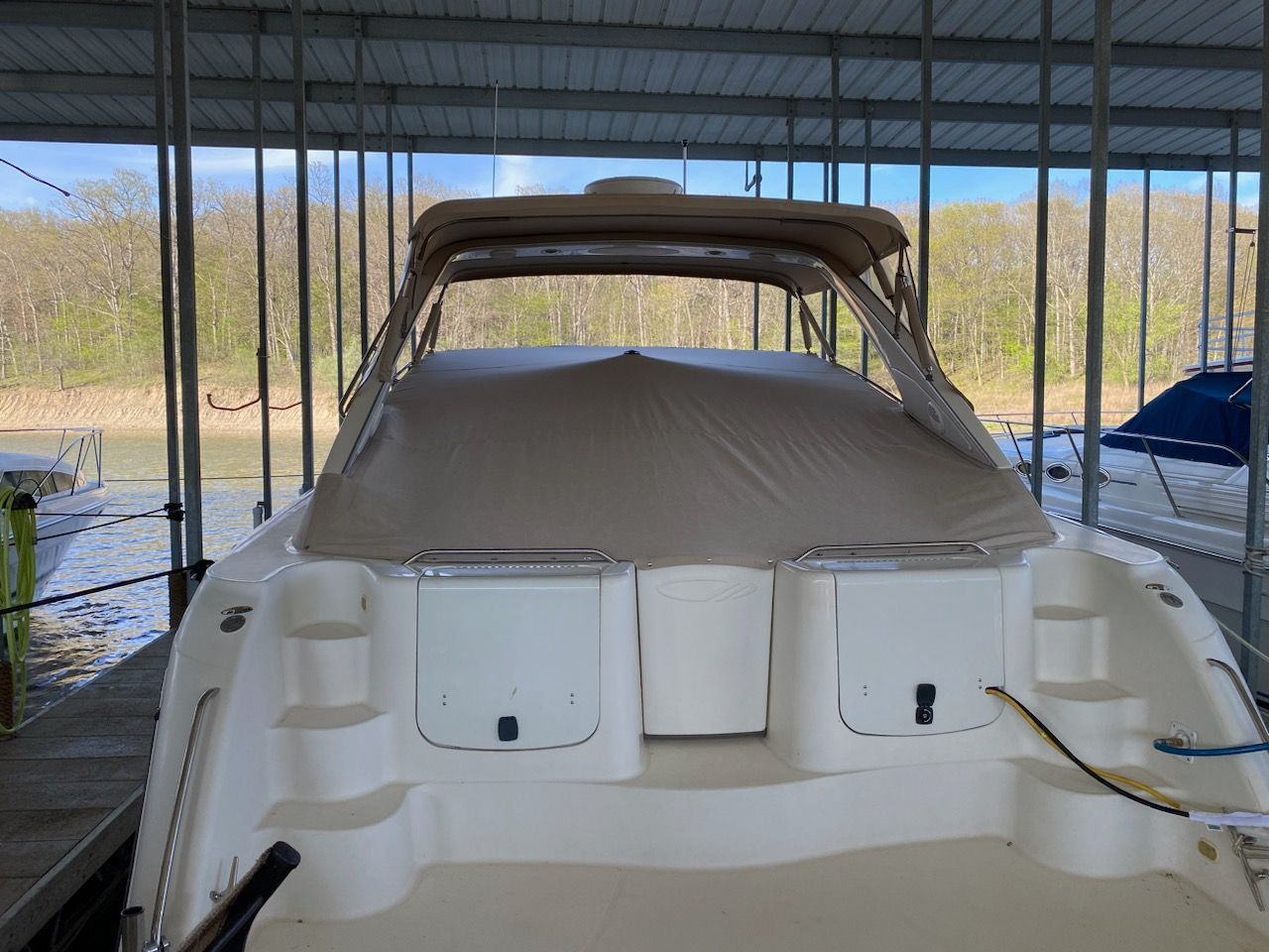 A beige boat under a dock cover with storage compartments, water, and trees in the background.