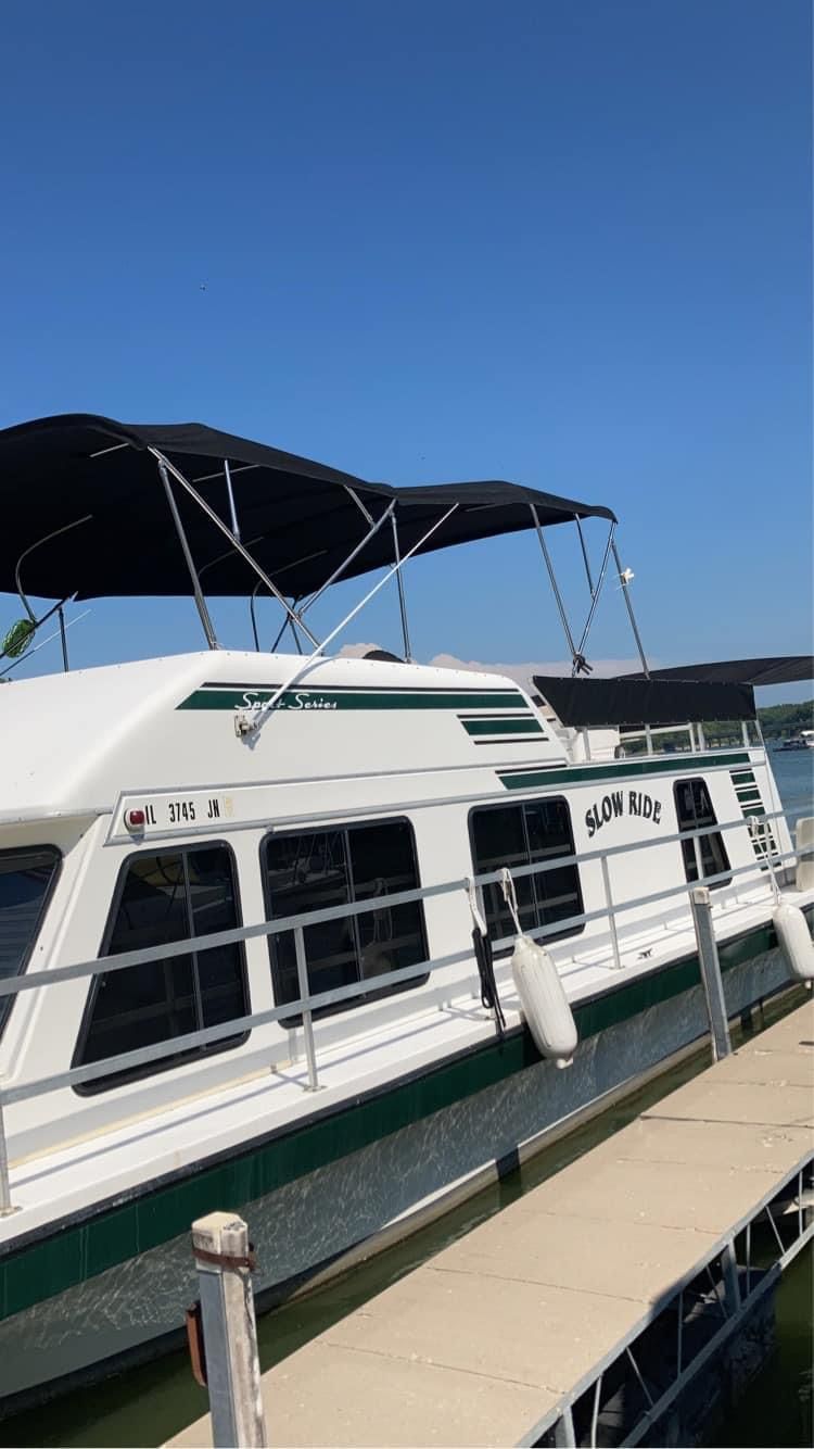 White and green houseboat docked on a sunny day, blue sky overhead. Black canopy on top.