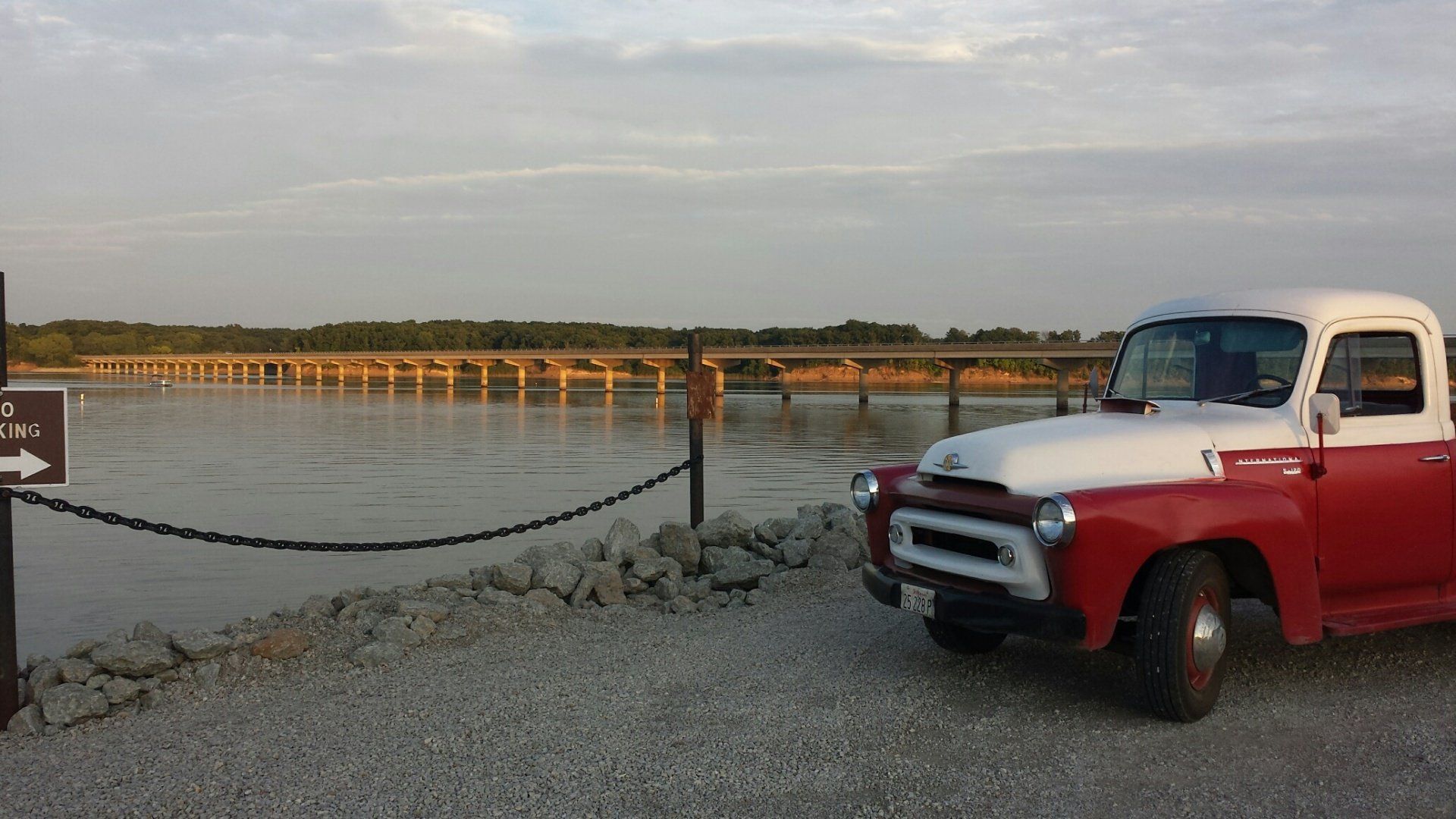 Red and white vintage pickup truck parked by river, bridge in background, sunny day.