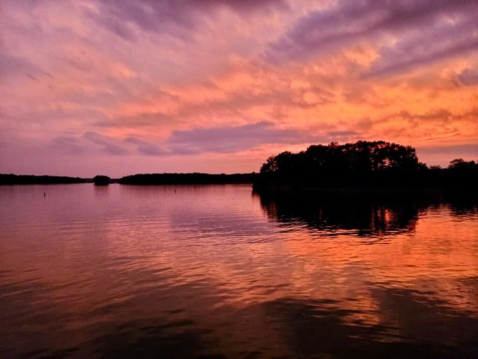 Sunset over a calm lake with vibrant orange and purple sky reflecting in the water. Silhouetted trees on the horizon.