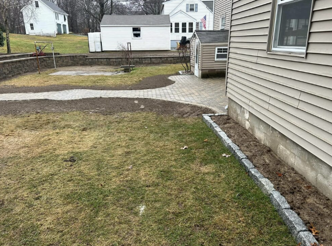Backyard with grass, a paved patio, retaining wall, and house.
