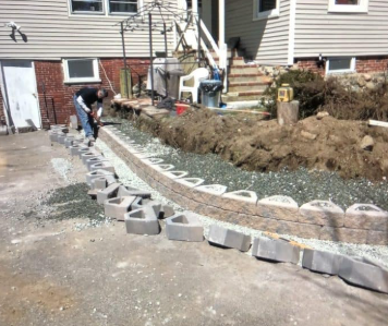 Man building a retaining wall with blocks along a driveway next to a house.
