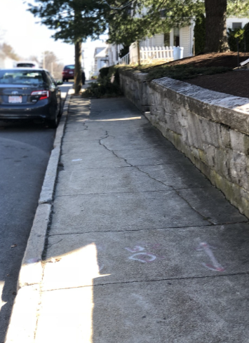 Sidewalk with cracks, bordered by a stone wall on the right and a parked car on the left.