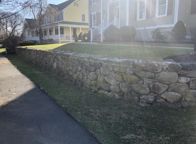 Stone retaining wall along a sidewalk, with grass and houses in the background. Bright sunlight.