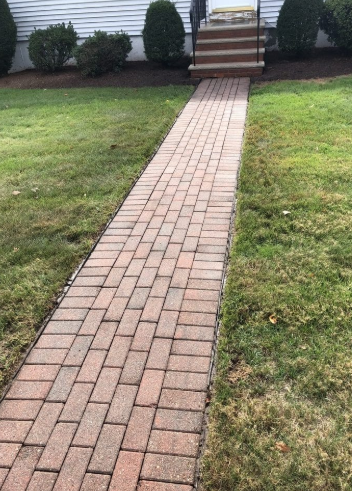 Brick path leads to steps in front of a house, surrounded by grass and bushes.