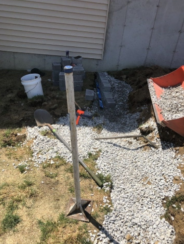 Construction site: Foundation being built with gray blocks. Gravel pathway, tools, bucket, and wheelbarrow visible.