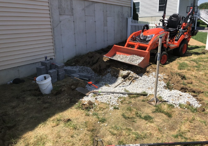Orange tractor with front loader digging gravel next to a house foundation, on a sunny day.