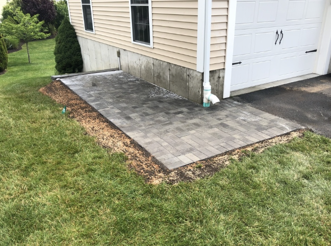 Brick patio next to a garage door, on a slight slope, with green grass.