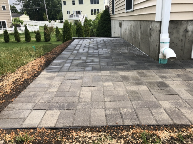 Paved patio beside a light-colored house. Brick pavers are grey and rectangular.