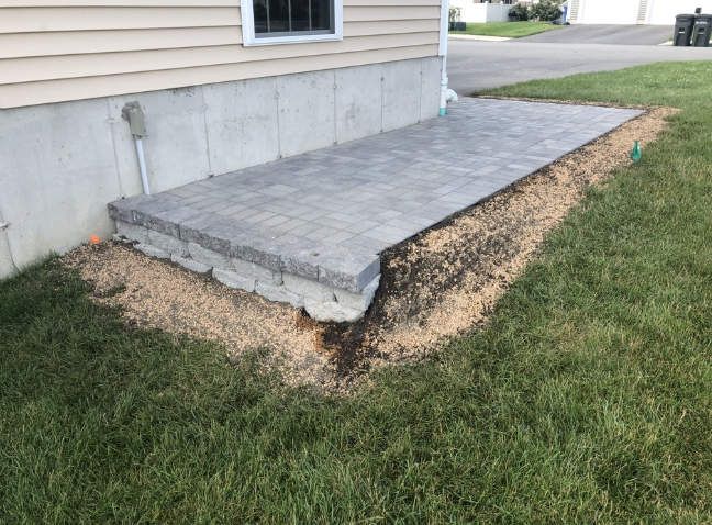 A small patio made of gray pavers next to a light-colored building with a grass yard.