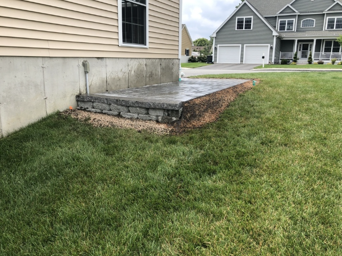 Concrete patio next to a house with a stone retaining wall and green grass.