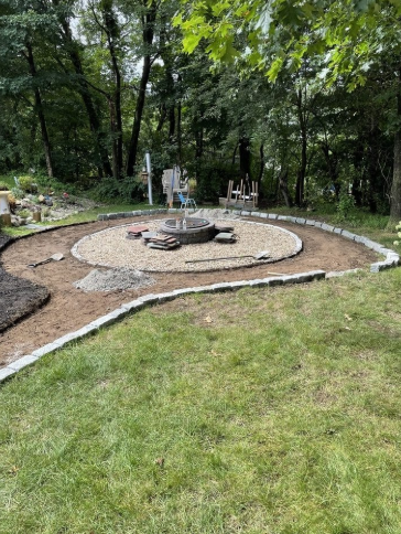 A partially constructed fire pit area in a grassy backyard, surrounded by a border and gravel.