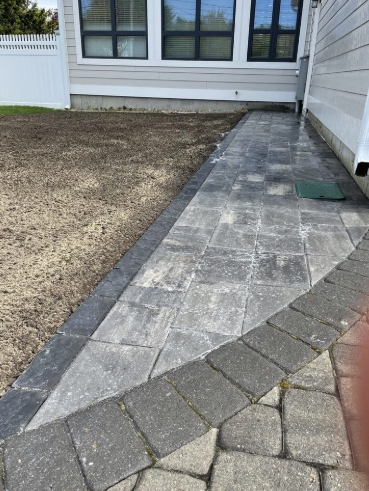 Brick walkway alongside a light-colored house. Pathway transitions to gravel.