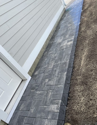 Dark gray brick pathway beside a light gray building with a white door and trim.