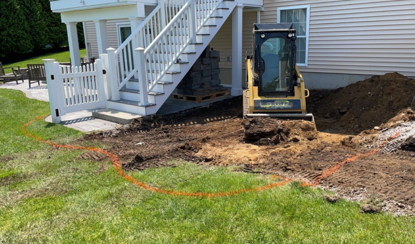 A skid steer excavating dirt near a white deck and stairs, outlined with orange paint on the grass.