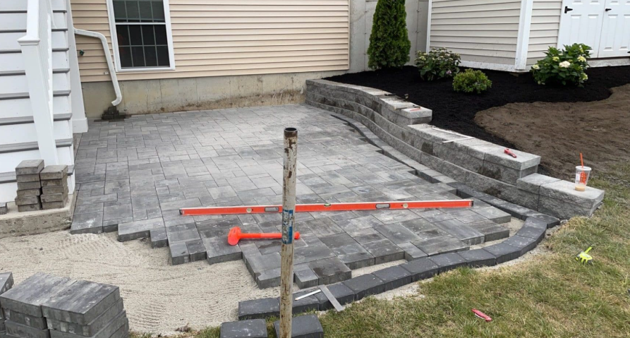 Patio construction: gray pavers, retaining wall, and leveling tools are in progress outdoors.