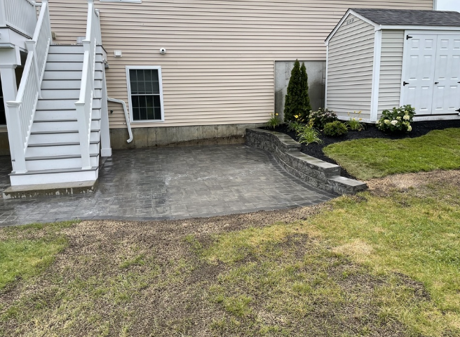 Backyard patio next to a house with stairs and a small shed; a retaining wall separates the patio from the lawn.