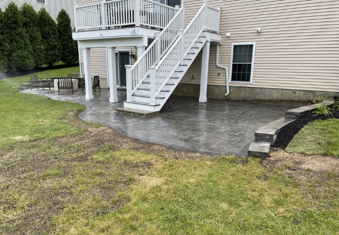 Backyard patio with stairs, deck, and retaining wall. Gray pavers on the ground. Green grass surrounds.