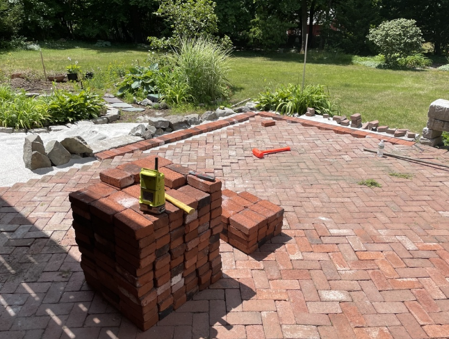 Brick patio under construction, with stack of bricks, level, and tools in a yard.