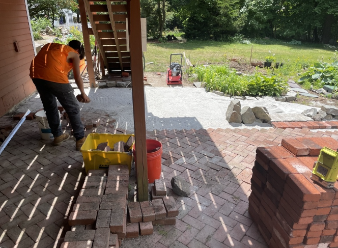Man laying brick pavers under a wooden staircase.  Orange shirt, yellow bucket, bricks and a compactor.