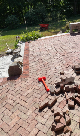 Brick patio construction; red bricks, orange mallet, wheelbarrow in a grassy yard.