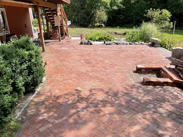 Red brick patio with a small brick fire pit, leading to a grassy yard with trees.