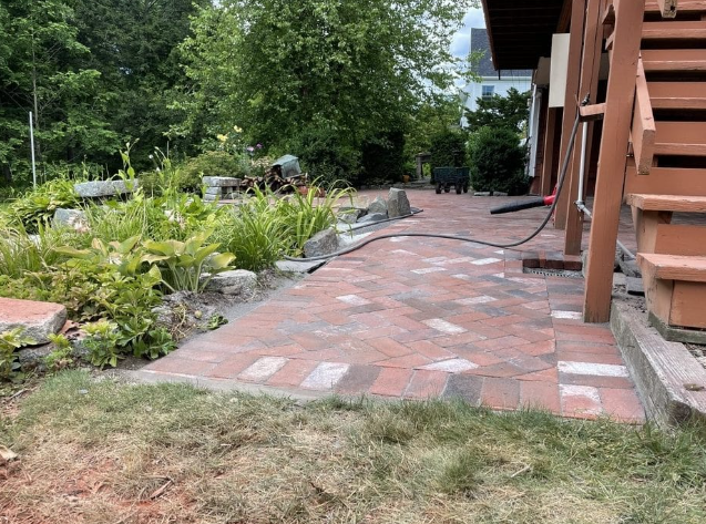 Brick patio next to a wooden structure, bordering a garden bed with green foliage.