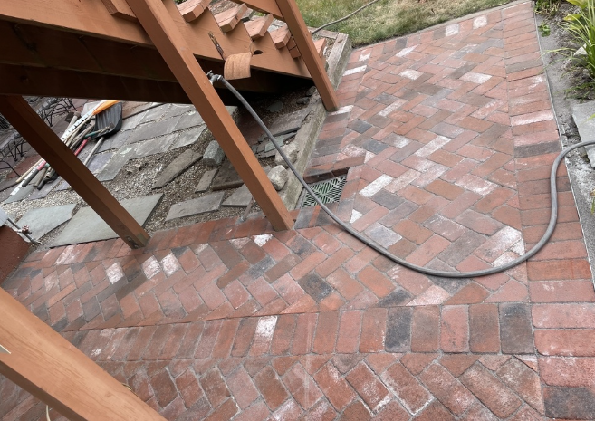 Brick patio with herringbone pattern under wooden deck stairs; a hose rests on the bricks.