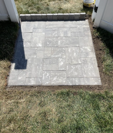 Gray pavers forming a small patio next to a white fence, surrounded by grass and dirt.