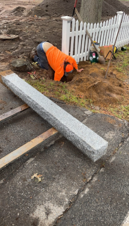 A person in an orange shirt kneels, working in a hole by a white picket fence, next to a large stone block on pavement.