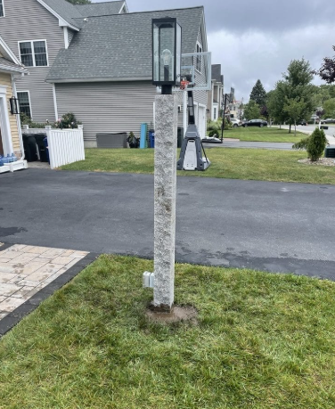 Stone light post in a grassy yard, with a dark light fixture at the top and a paved driveway behind.
