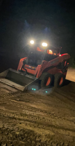 Orange skid steer loader working at night, headlights on, scooping sand or dirt.