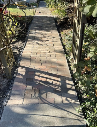 Brick pathway leading uphill through a garden, bordered by plants and trellises.