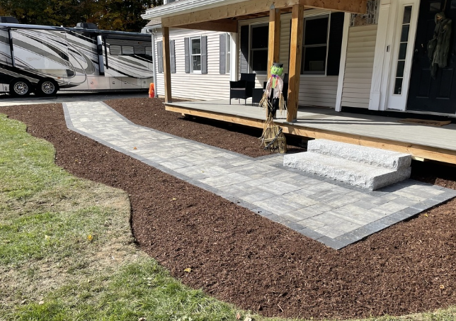 Brick pathway leading to a porch with steps; surrounded by brown mulch and green grass; a camper is in the background.