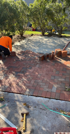 A person laying red bricks in a herringbone pattern on a patio; trees and equipment visible in the background.