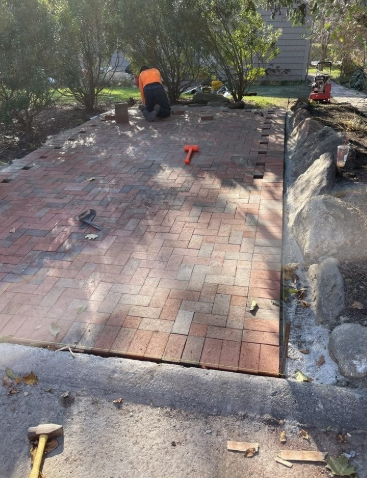 Person laying brick pavement outdoors. Red bricks, orange safety vest, tools visible, trees in the background.