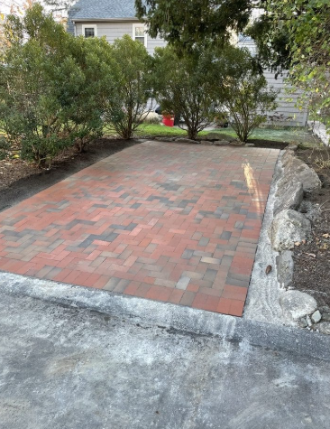 Brick patio with a concrete border, beside a rock formation and driveway, shrubs in the background.