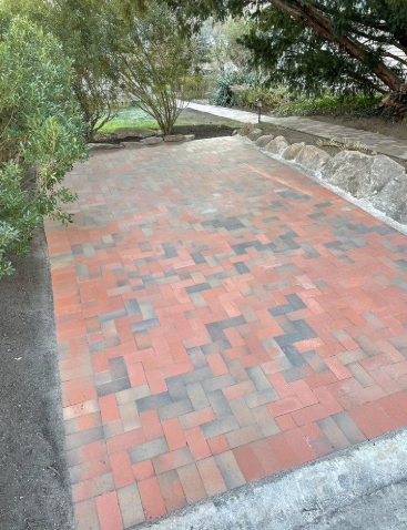 Brick paver driveway with red, grey, and black bricks, bordered by soil and a sidewalk, in a garden setting.