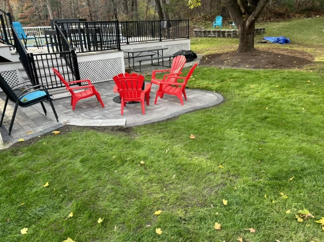 Backyard patio with red chairs, fire pit, and a deck. Green lawn surrounds the space.