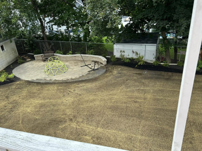 Backyard with freshly seeded lawn, concrete play area, a small white shed, and surrounding trees.