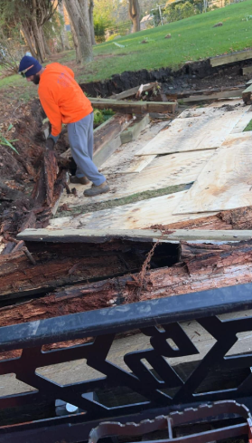 Man in orange shirt working on damaged wooden structure outdoors. Green grass and trees in background.