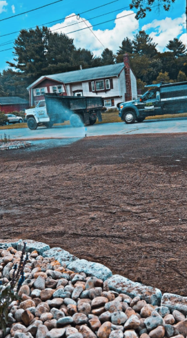 Trucks spraying water on freshly laid dirt in front of a house, blue sky above.