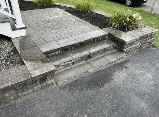 Stone steps leading to a paved patio, bordered by low retaining walls and planters with green plants.