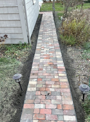 Brick path bordered by soil and grass, leading towards a green lawn, two solar lights.