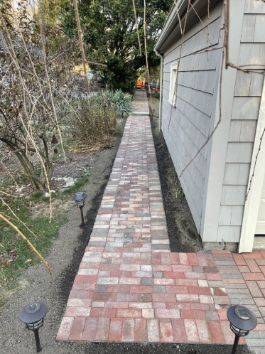 Brick pathway alongside a gray building, leading into a yard with greenery. Two solar lights.
